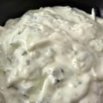 Close-up of a bowl of homemade tzatziki sauce, garnished with fresh dill and a drizzle of olive oil, placed on a wooden table next to sliced cucumbers and pita bread.