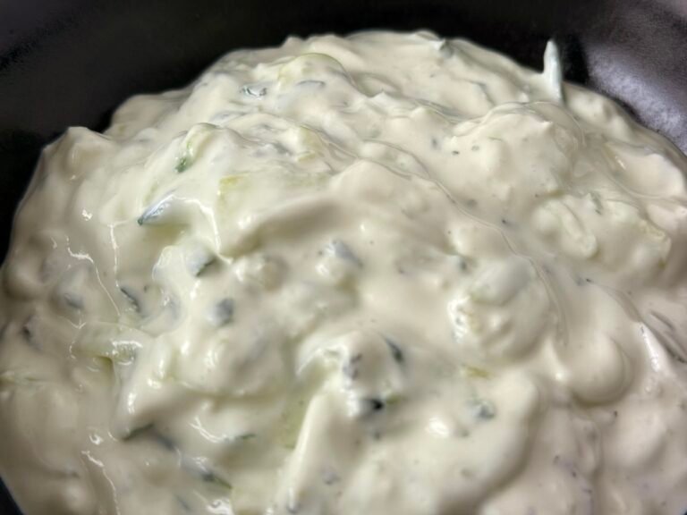 Close-up of a bowl of homemade tzatziki sauce, garnished with fresh dill and a drizzle of olive oil, placed on a wooden table next to sliced cucumbers and pita bread.