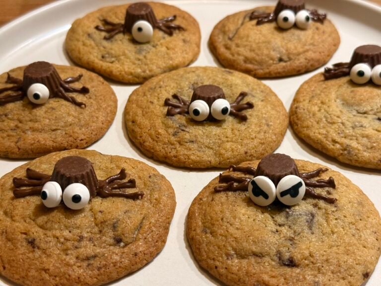 A plate of Halloween-themed spider cookies topped with mini peanut butter cups as spider bodies, chocolate icing legs, and candy eyes. These chewy cookies are dotted with dark chocolate chunks, adding a festive, spooky touch to any Halloween dessert table.