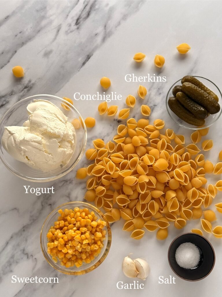 Conchiglie pasta, plain yoghurt, chopped gherkins, sweetcorn, minced garlic, and salt on a wooden board. Essential ingredients for a versatile pasta salad.