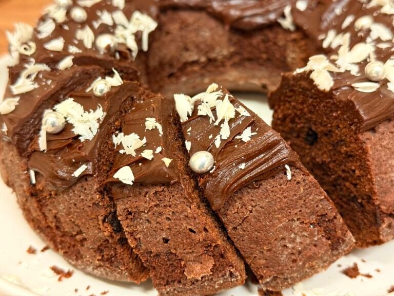 Rich chocolate bundt cake draped in glossy dark chocolate ganache and topped with delicate shavings of white chocolate, sliced and ready to serve.