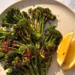 Charred grilled broccoli served with lemon wedges and red pepper flakes on a rustic plate